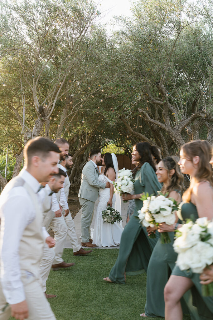 Bride and groom kissing while wedding party walking in front of them the wedding party is slightly blurred while the couple is in focus 