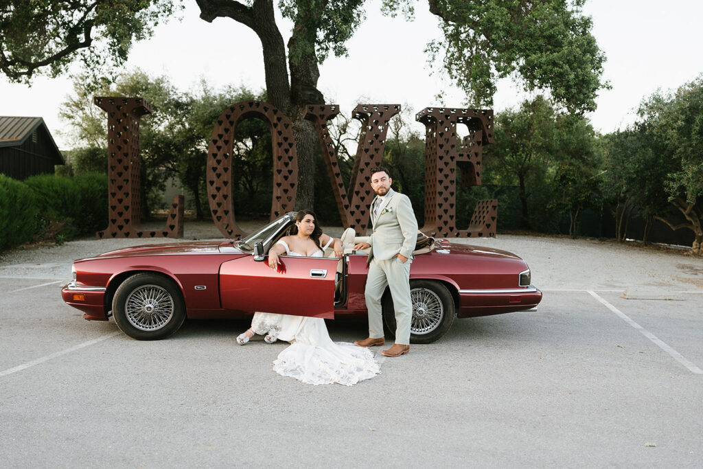 Bride and groom in red vintage jaguar. Bride sitting in the drivers set with door open while groom is leaned against the side of the door
