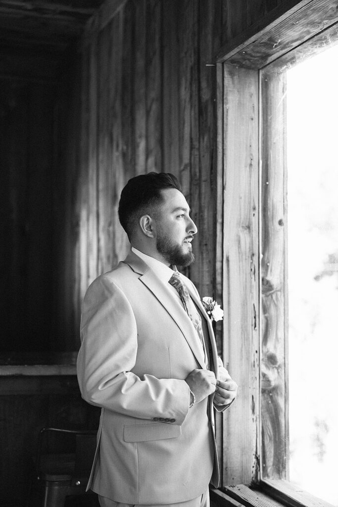Black and white photo of a groom standing in front of a window buttoning his suit