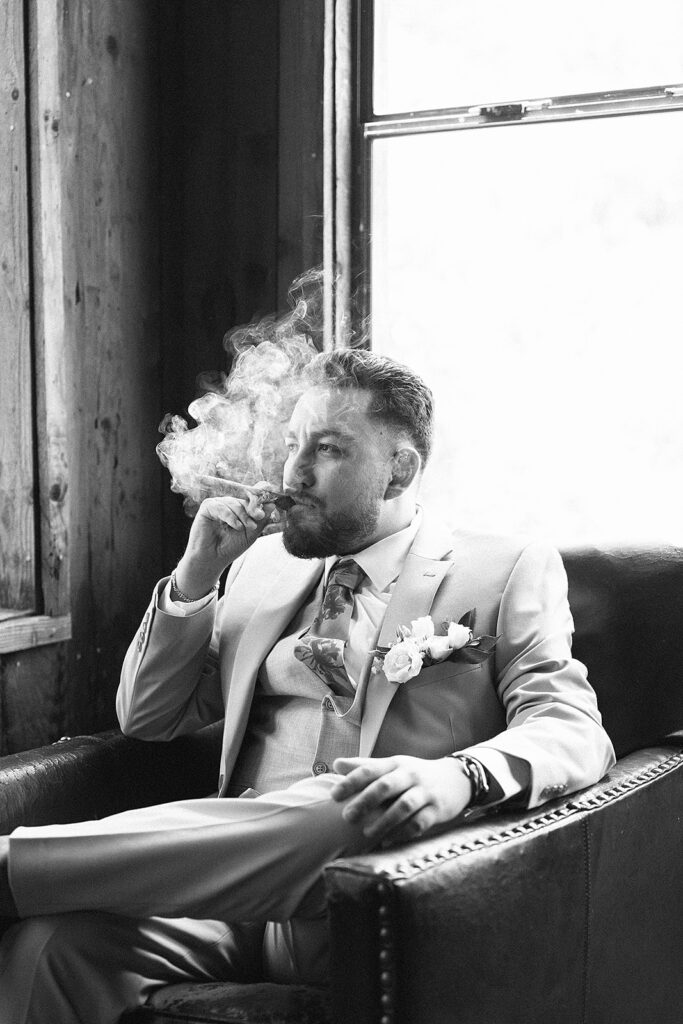 Black and white photo of a groom sitting on a leather chair smoking a cigar